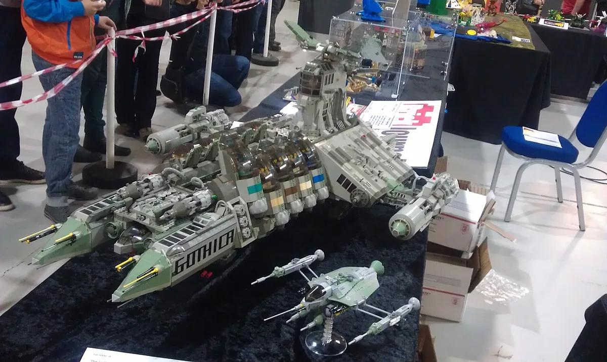 Een tentoonstellingshal met lange tafels waarop LEGO-modellen zijn uitgestald, met bezoekers die erlangs lopen.