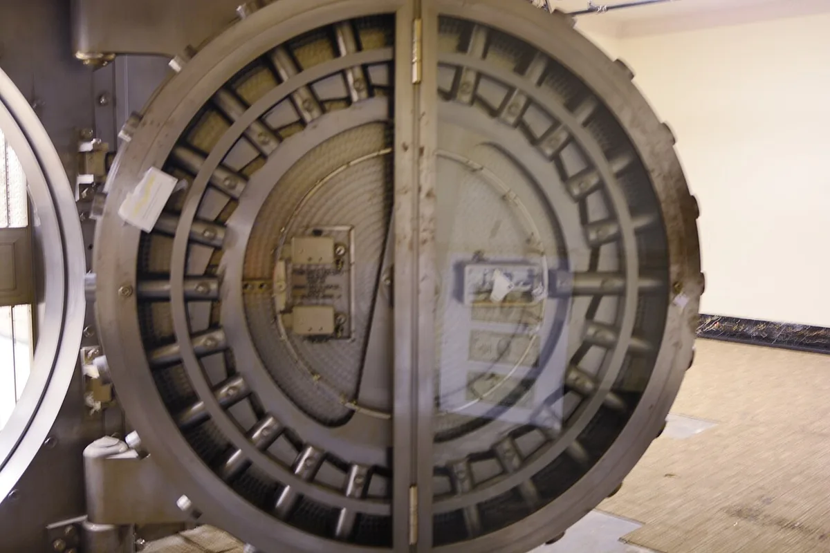 Een zware ronde kluisdeur van staal staat open in een lege ruimte, met zichtbare grendels langs de rand.